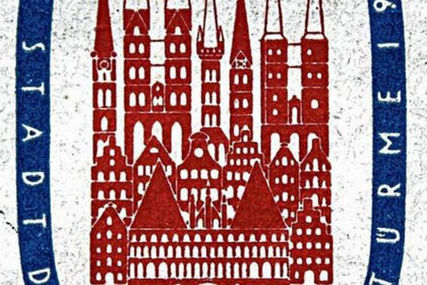 Man sieht den Ausschnitt der Briefmarke von 1956. In der Mitte sieht man in roter Farbe Dächer von Lübeck, sowie die Kirchen und das Holstentor. Umrandet sind diese Häuser von einem blauen breiten Ring, auf dem in weißer Schrift: Stadt und Türme aufgedruckt sind - Copyright: Stadtarchiv Hansestadt Lübeck
